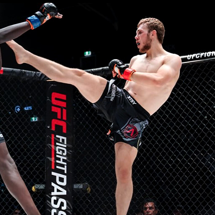 Krane Burke throwing a high kick inside the cage, his opponent's leg raised to block.