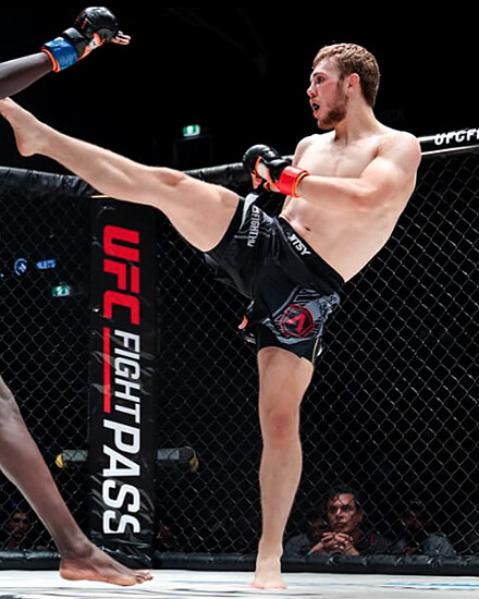 Krane Burke throwing a high kick inside the cage, his opponent's leg raised to block.